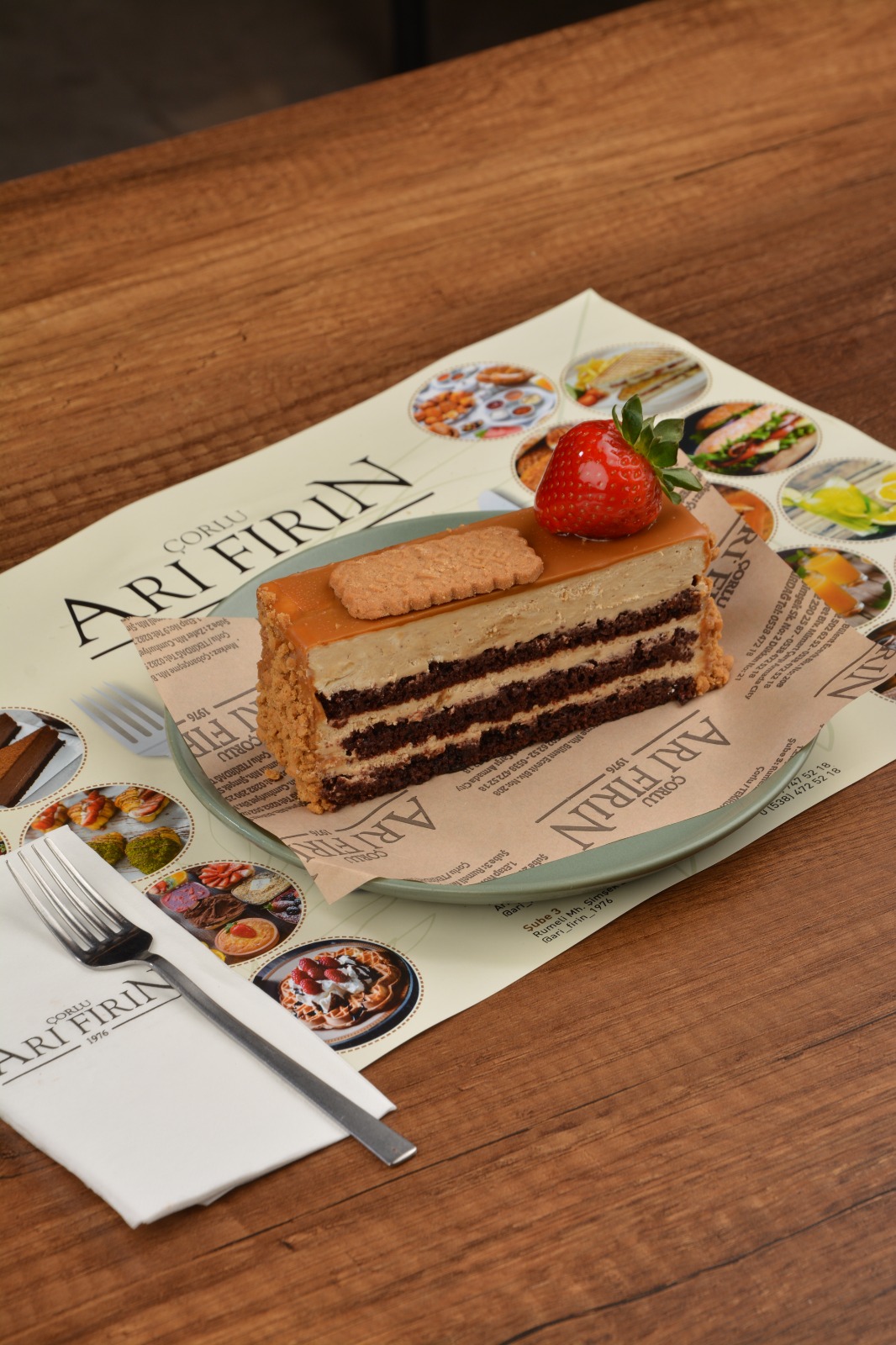 Kakaolu pandispanya katmanları arasında yoğun krema, üzerinde parlak karamel sos, taze çilek ve karamelize bisküvi bulunan nefis dilim pasta. – Arı Fırın Çorlu